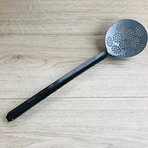 Puede incluir: Una cuchara ranurada de metal negro con un mango largo. La cuchara tiene una cabeza circular con un patrón de pequeños agujeros.