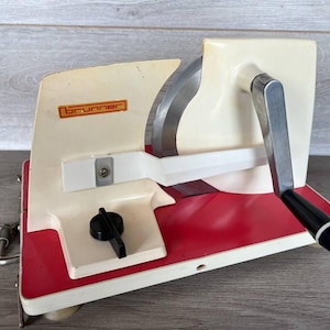 May include: A vintage Brunner meat slicer in cream and red. It has a large, curved blade, a black handle, and a metal clamp. The Brunner logo is displayed on the side of the appliance.