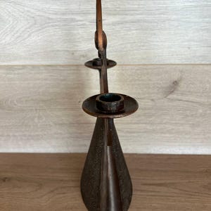 May include: A bronze-colored candle holder with a tall, slender design. The holder features a conical base, a central stem, and two circular platforms to hold candles. The metal has a textured surface, adding visual interest to the piece.