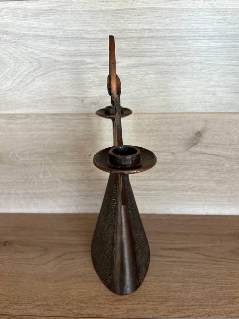 May include: A bronze-colored candle holder with a tall, slender design. The holder features a conical base, a central stem, and two circular platforms to hold candles. The metal has a textured surface, adding visual interest to the piece.