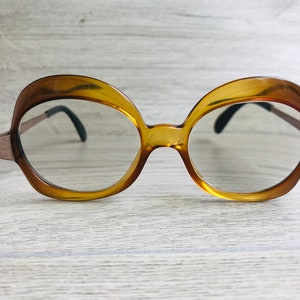 Puede incluir: Un par de gafas con montura de plástico marrón y lentes redondas. Las gafas tienen una forma única y curvada.