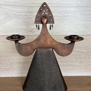 May include: A copper-toned candle holder with a stylized figure design. The figure has a triangular head with decorative holes, outstretched arms holding candle cups, and a textured, cone-shaped base. The candle holder is a decorative home accent.