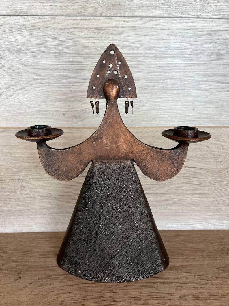 May include: A copper-toned candle holder with a stylized figure design. The figure has a triangular head with decorative holes, outstretched arms holding candle cups, and a textured, cone-shaped base. The candle holder is a decorative home accent.