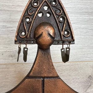 May include: A bronze-colored, decorative wall hanging with a triangular top featuring ornate scrollwork and circular cutouts. Below the triangle is a rounded shape, and small metal pieces hang from the bottom. The piece has a rustic, vintage aesthetic.