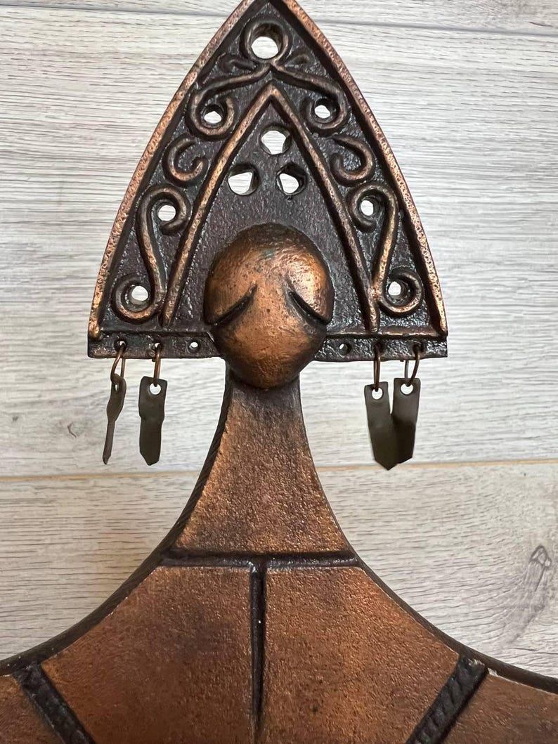 May include: A bronze-colored, decorative wall hanging with a triangular top featuring ornate scrollwork and circular cutouts. Below the triangle is a rounded shape, and small metal pieces hang from the bottom. The piece has a rustic, vintage aesthetic.