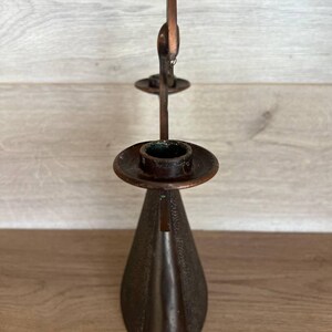 May include: A vintage bronze-colored candle holder with a unique design. The candle holder has a conical base, a circular dish to hold the candle, and a decorative element at the top. The metal has a textured appearance.