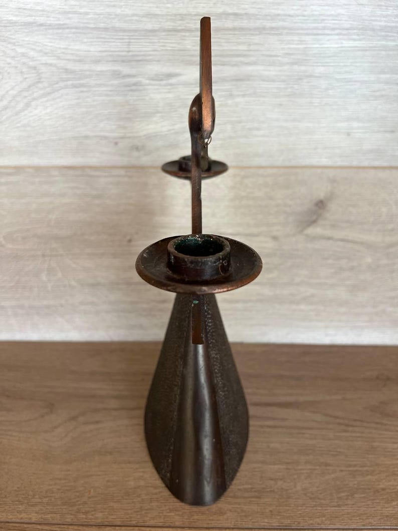 May include: A vintage bronze-colored candle holder with a unique design. The candle holder has a conical base, a circular dish to hold the candle, and a decorative element at the top. The metal has a textured appearance.