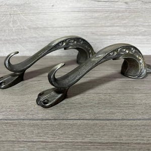May include: Two antique silver-toned metal door handles with ornate designs. The handles are curved and have a hook-like shape at the end.