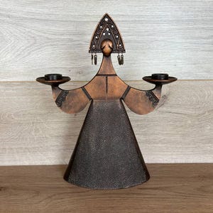 May include: A bronze-colored metal candle holder in the shape of a stylized figure. The figure has outstretched arms holding circular candle platforms. The head is adorned with a decorative headdress. The base of the figure is a flared cone shape.