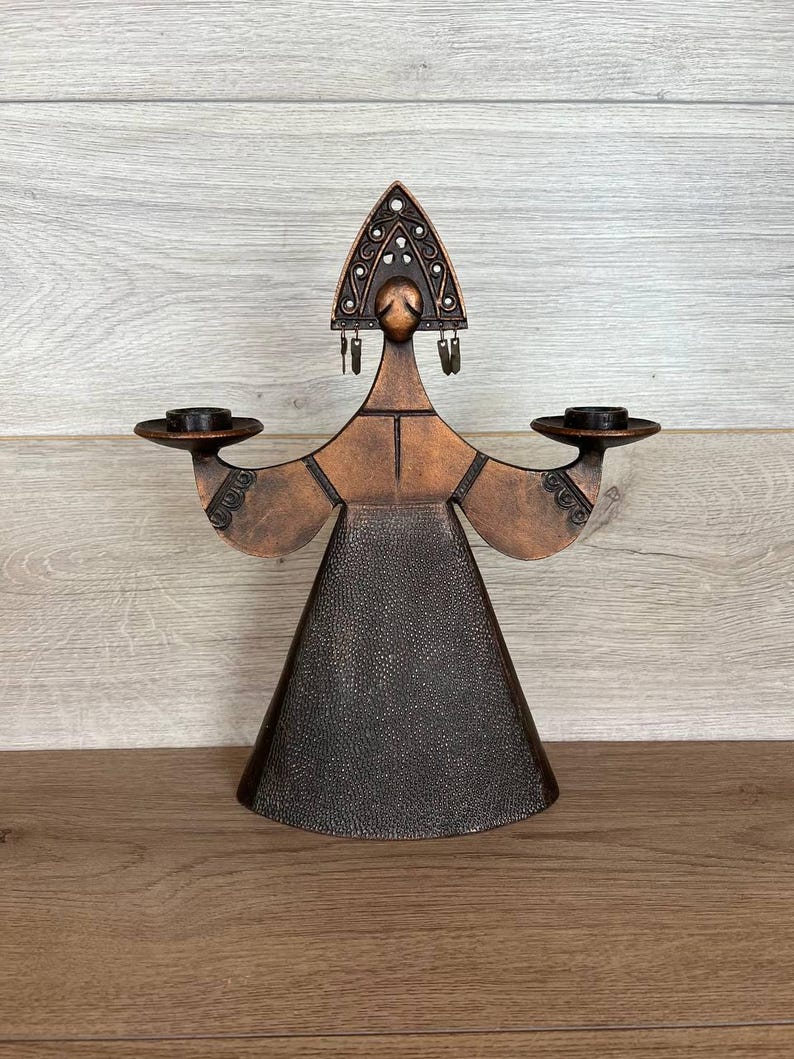 May include: A bronze-colored metal candle holder in the shape of a stylized figure. The figure has outstretched arms holding circular candle platforms. The head is adorned with a decorative headdress. The base of the figure is a flared cone shape.