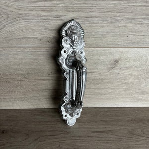 Może przedstawiać: Srebrny klamka do drzwi z ozdobnym wzorem przedstawiającym twarz i motywy kwiatowe.