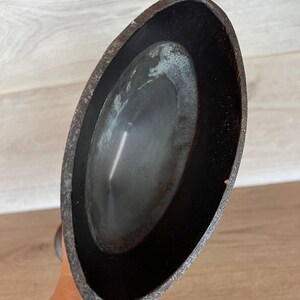 May include: A close-up view of a dark, oval-shaped metal object. The object has a textured, aged appearance with a dark interior and a slightly lighter, weathered exterior. The object is held by a hand.