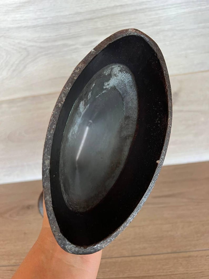May include: A close-up view of a dark, oval-shaped metal object. The object has a textured, aged appearance with a dark interior and a slightly lighter, weathered exterior. The object is held by a hand.