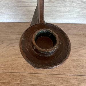 May include: A close-up of a brown metal candlestick holder. The candlestick holder has a circular base with a central opening for a candle. The stem is long and curved, with a textured surface.