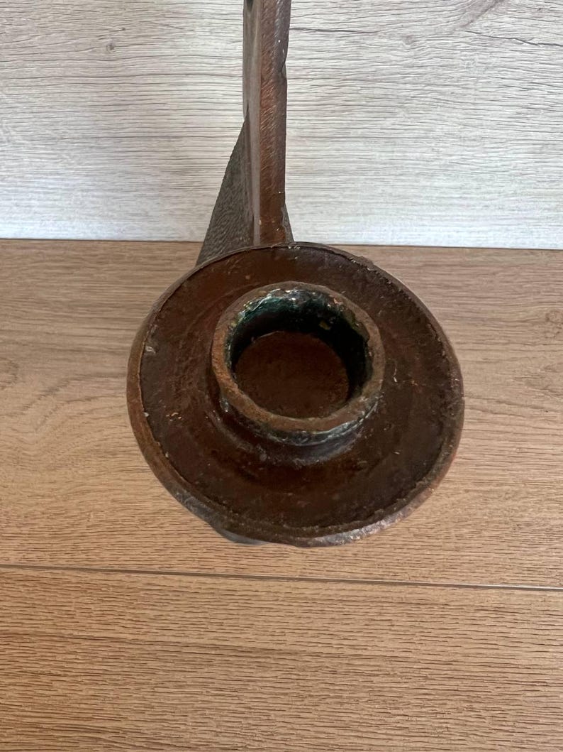 May include: A close-up of a brown metal candlestick holder. The candlestick holder has a circular base with a central opening for a candle. The stem is long and curved, with a textured surface.