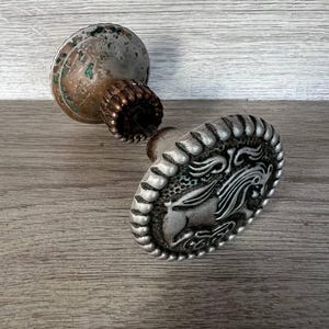 May include: A silver-toned metal door knob featuring an oval face with a raised relief design of a horse head. The knob has a decorative fluted collar and a weathered, tarnished circular base with visible green oxidation and copper-coloured patches.