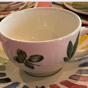 Puede incluir: Un juego de taza de t&eacute; y plato a juego de cer&aacute;mica blanca con un dise&ntilde;o pintado a mano de hojas verdes en la taza y un borde colorido de pinceladas cortas y gruesas en varios tonos, incluyendo azul, amarillo, rojo y verde en el plato.