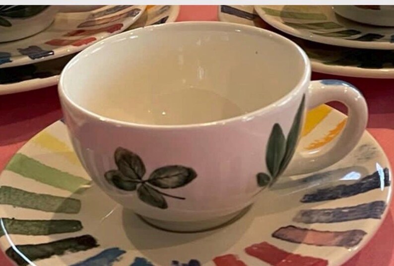 Puede incluir: Un juego de taza de t&eacute; y plato a juego de cer&aacute;mica blanca con un dise&ntilde;o pintado a mano de hojas verdes en la taza y un borde colorido de pinceladas cortas y gruesas en varios tonos, incluyendo azul, amarillo, rojo y verde en el plato.