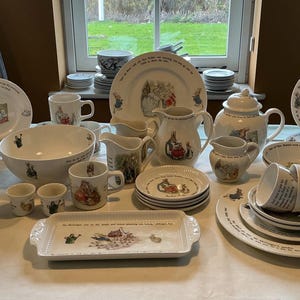 May include: A full white ceramic Peter Rabbit tea set. The set includes plates, cups, saucers, mugs, a teapot, creamer, sugar bowl, and serving dishes. Each piece features illustrations of Peter Rabbit and other characters.