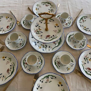 May include: A white ceramic cake stand with a gold handle and a floral design. The stand is filled with white ceramic plates and cups with a blue trim and floral designs.