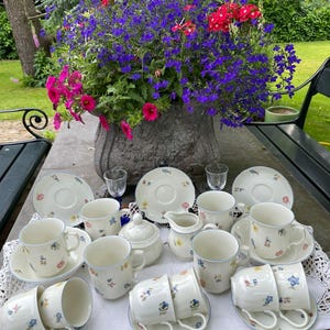 Pode incluir: Um conjunto de chá com desenhos florais, incluindo xícaras, pires, açucareiro e cremeira. O conjunto está disposto sobre uma mesa, com um vaso de flores cheio de flores roxas, rosas e vermelhas.