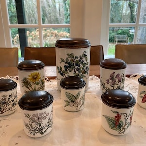 May include: Eight ceramic canisters with brown lids and floral designs. The canisters are arranged in a circle on a white surface. The designs include berries, flowers, and leaves.
