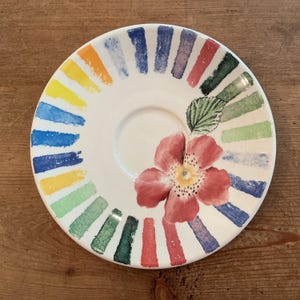 Puede incluir: Un platillo redondo de cer&aacute;mica blanca con un borde decorativo de pinceladas rectangulares multicolores en tonos azul, amarillo, naranja, rojo y verde. Presenta una gran flor rosa con el centro amarillo y una hoja verde pintada.