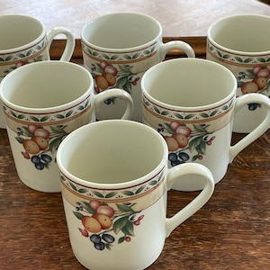 May include: Six white ceramic mugs with a decorative band featuring fruit and foliage. The mugs have a curved handle and a cream-colored interior. The mugs are arranged on a wooden surface.