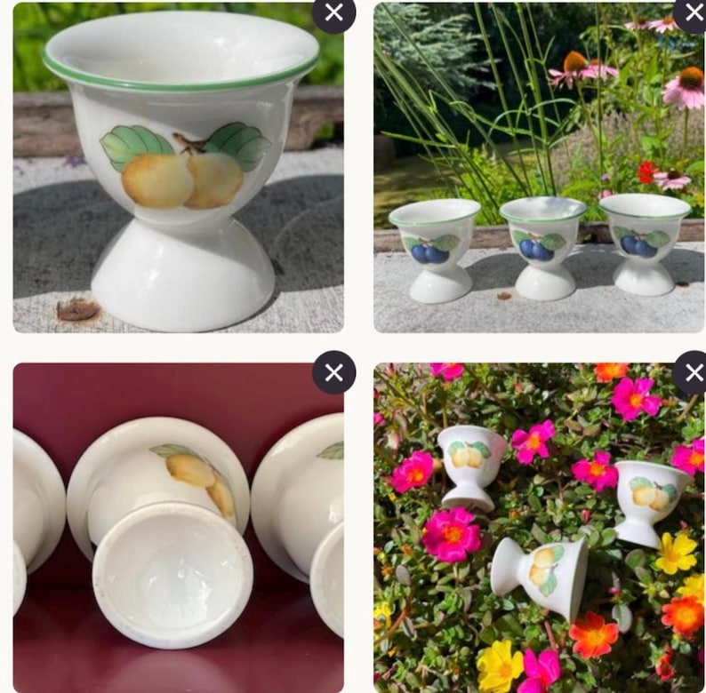 May include: A set of white ceramic egg cups featuring a flared rim with a thin green border. Each cup is decorated with a painted illustration of various fruits, including yellow pears and dark blue plums, centred on the exterior surface.
