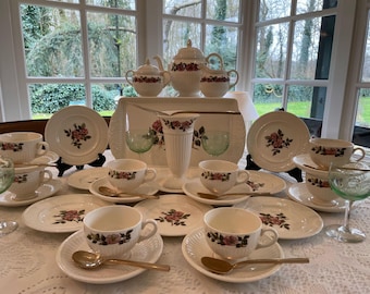 May include: A vintage tea set featuring a floral rose design. The set includes teacups, saucers, plates, a teapot, sugar bowl, creamer, and a serving tray. The set is white with pink and red roses and green leaves.