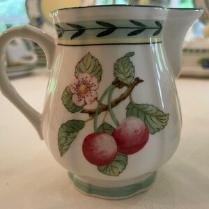 May include: A white ceramic cream jug featuring a painted botanical motif of two red cherries, green leaves, and a small white flower. The rim and base are decorated with a thin green leaf pattern and a yellow band, with a curved handle.