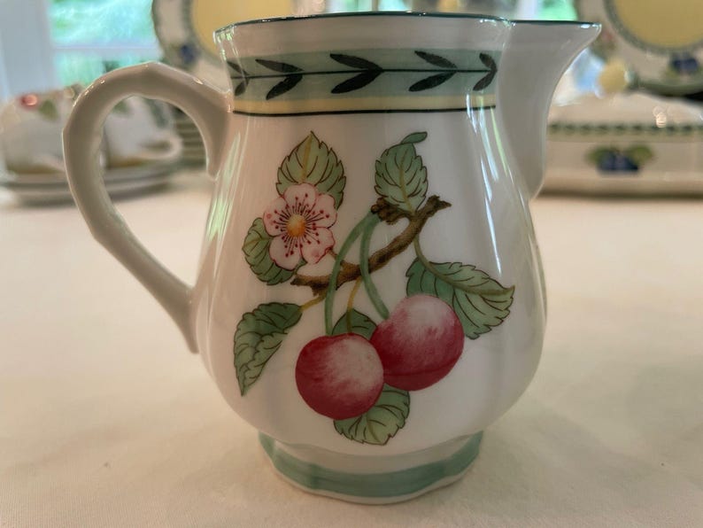May include: A white ceramic cream jug featuring a painted botanical motif of two red cherries, green leaves, and a small white flower. The rim and base are decorated with a thin green leaf pattern and a yellow band, with a curved handle.