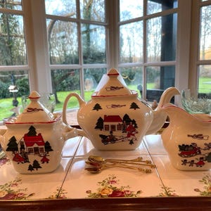 May include: A white ceramic tea set with a winter village design. The set includes a teapot, creamer, and sugar bowl. Each piece features a red roofed house, trees, and figures in a snowy landscape.