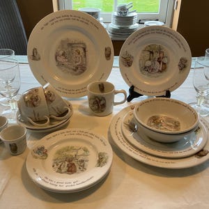May include: A collection of white ceramic tableware featuring illustrations and text. The set includes plates, bowls, cups, and a mug. The illustrations depict scenes with characters. Clear stemmed glasses are also present.