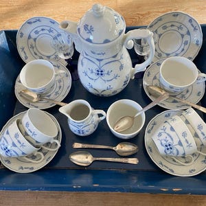 Puede incluir: Un juego de té completo con un diseño floral blanco y azul, que incluye una tetera, tazas, platillos, cucharas, una jarra de crema y un azucarero. El juego está dispuesto sobre una bandeja de madera azul.