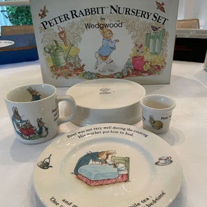 May include: A Wedgwood Peter Rabbit nursery set including a box, a mug, a small cup, and a plate. The plate features an illustration of a rabbit in bed with text reading, 'Peter was not very well during the evening. His mother put him to bed.'