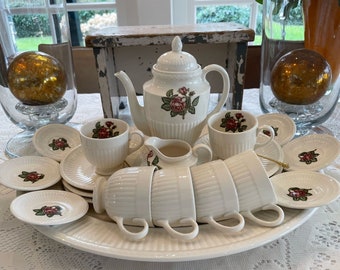 May include: A white teapot with a floral design and six white teacups with floral designs on a white tablecloth. The teapot and teacups are arranged in a circle around a white serving tray.