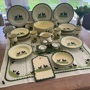 Puede incluir: Una colección de vajilla de cerámica color crema con borde festoneado verde y motivo de gallo. Incluye platos, cuencos, tazas, una jarra, saleros y pimenteros, y candelabros. Una esterilla y una tabla de cortar a juego completan el conjunto.