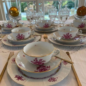 Könnte beinhalten: Ein Set aus sechs weißen Teetassen und Untertassen mit einem rosafarbenen Blumenmuster. Die Tassen und Untertassen sind auf einer weißen Tischdecke mit goldenem Besteck angeordnet.