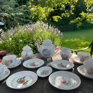 May include: A white porcelain tea set featuring Peter Rabbit illustrations, including a teapot, a lidded sugar bowl, a creamer, four teacups with matching saucers, and three small plates. Each piece displays colorful character scenes.