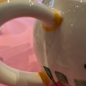 Puede incluir: Una tetera de cer&aacute;mica blanca con un asa curva que presenta detalles pintados en amarillo brillante en los puntos de uni&oacute;n. El cuerpo de la tetera muestra un dise&ntilde;o floral parcial con hojas verdes y p&eacute;talos rosas visibles en la parte inferior derecha.