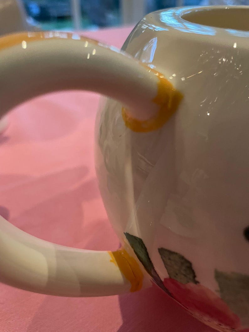 Puede incluir: Una tetera de cer&aacute;mica blanca con un asa curva que presenta detalles pintados en amarillo brillante en los puntos de uni&oacute;n. El cuerpo de la tetera muestra un dise&ntilde;o floral parcial con hojas verdes y p&eacute;talos rosas visibles en la parte inferior derecha.