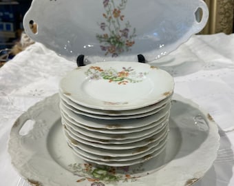 May include: A set of vintage white porcelain plates with floral designs. The set includes a large oval serving platter with handles, a smaller round plate, and a stack of smaller round plates.
