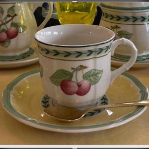 May include: A white ceramic teacup and saucer set featuring a painted cherry motif with green leaves. The saucer has a pale yellow centre and a green rim. A small silver-toned metal spoon rests on the saucer. The cup has a scalloped edge and a handle.