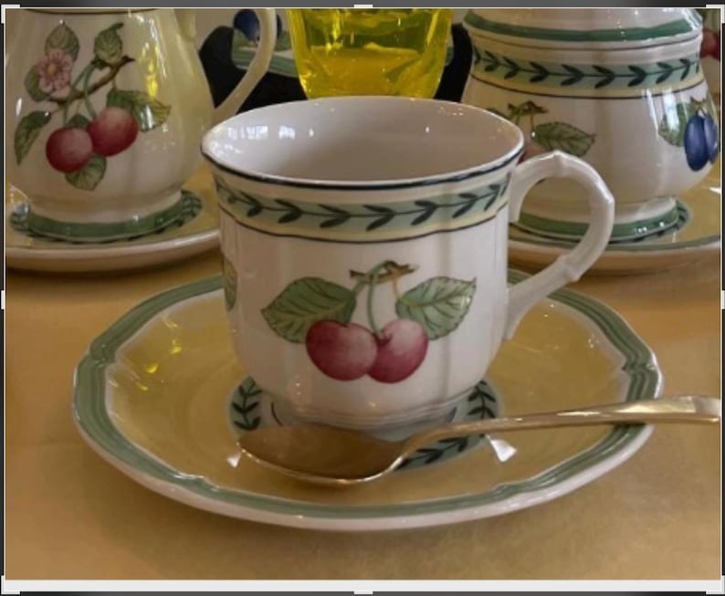 May include: A white ceramic teacup and saucer set featuring a painted cherry motif with green leaves. The saucer has a pale yellow centre and a green rim. A small silver-toned metal spoon rests on the saucer. The cup has a scalloped edge and a handle.