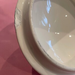 Puede incluir: Una tapa de cer&aacute;mica blanca de forma circular con un acabado liso y brillante. El borde de la tapa presenta una muesca visible en el material cer&aacute;mico, revelando la textura interior blanca de la pieza. La parte inferior es c&oacute;ncava y lisa.