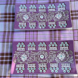 Op de afbeelding: Twee rechthoekige, paarse decoratieve trays met witte en teal accenten. Elke tray heeft een bloemen- en scrollontwerp. De trays liggen op een paars-wit geruite tafelkleed.