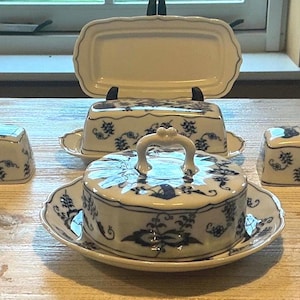 Può includere: Una collezione di burriere in ceramica bianca con un motivo floreale blu. Il set comprende quattro burriere rettangolari con coperchio e una burriera rotonda con manico centrale e coperchio abbinato sullo sfondo.