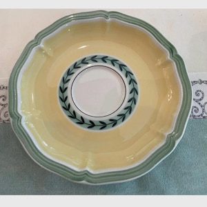 May include: A round ceramic saucer featuring a pale yellow centre and a scalloped rim accented with sage green bands. The inner circle displays a white base decorated with a dark green leaf wreath pattern, creating a distinct border around the centre.