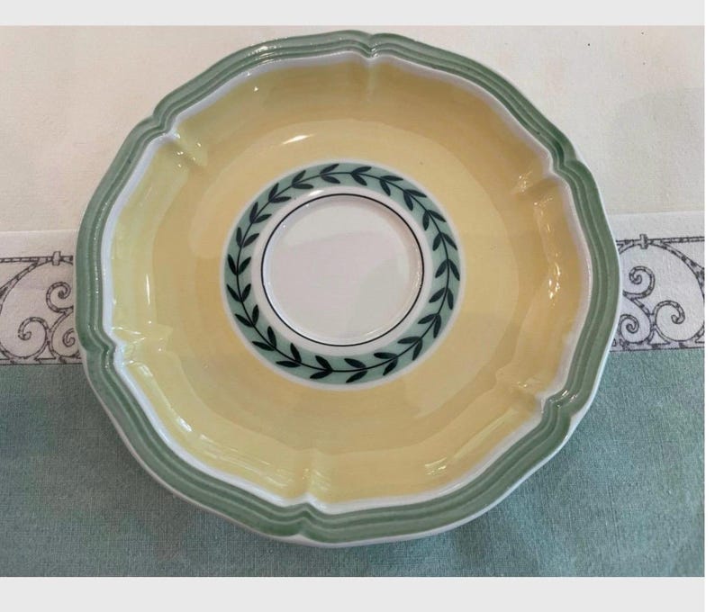 May include: A round ceramic saucer featuring a pale yellow centre and a scalloped rim accented with sage green bands. The inner circle displays a white base decorated with a dark green leaf wreath pattern, creating a distinct border around the centre.